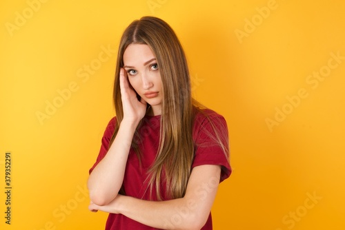 Very bored Beautiful Young beautiful caucasian girl wearing red t-shirt over isolated yellow background holding hand on cheek while support it with another crossed hand, looking tired and sick,