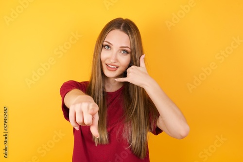 Beautiful Young beautiful caucasian girl wearing red t-shirt over isolated yellow background smiling cheerfully and pointing to camera while making a call you later gesture, talking on phone