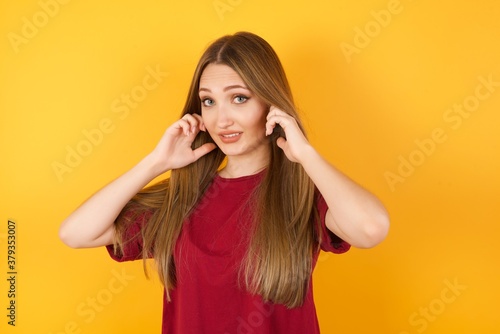 Beautiful Young beautiful caucasian girl wearing red t-shirt over isolated yellow background covering ears with fingers with annoyed expression for the noise of loud music. Deaf concept.