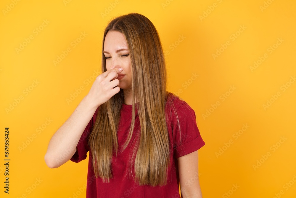Beautiful Young beautiful caucasian girl wearing red t-shirt over isolated yellow background smelling something stinky and disgusting, intolerable smell, holding breath with fingers on nose. Bad smell Beautiful Young beautiful caucasian girl wearing red t-shirt over isolated yellow background smelling something stinky and disgusting, intolerable smell, holding breath with fingers on nose. Bad smell