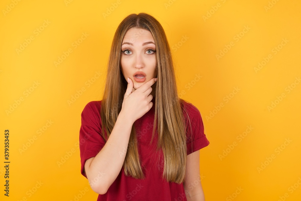 Beautiful Young beautiful caucasian girl wearing red t-shirt over isolated yellow background Looking fascinated with disbelief, surprise and amazed expression with hands on chin Beautiful Young beautiful caucasian girl wearing red t-shirt over isolated yellow background Looking fascinated with disbelief, surprise and amazed expression with hands on chin