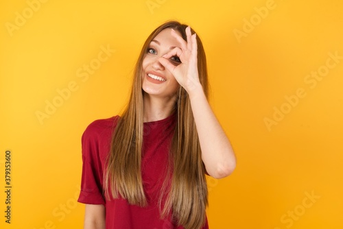 Beautiful Young beautiful caucasian girl wearing red t-shirt over isolated yellow background doing ok gesture with hand smiling, eye looking through fingers with happy face.