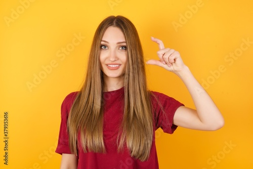 Beautiful Young beautiful caucasian girl wearing red t-shirt over isolated yellow smiling and confident gesturing with hand doing small size sign with fingers looking and the camera. Measure concept