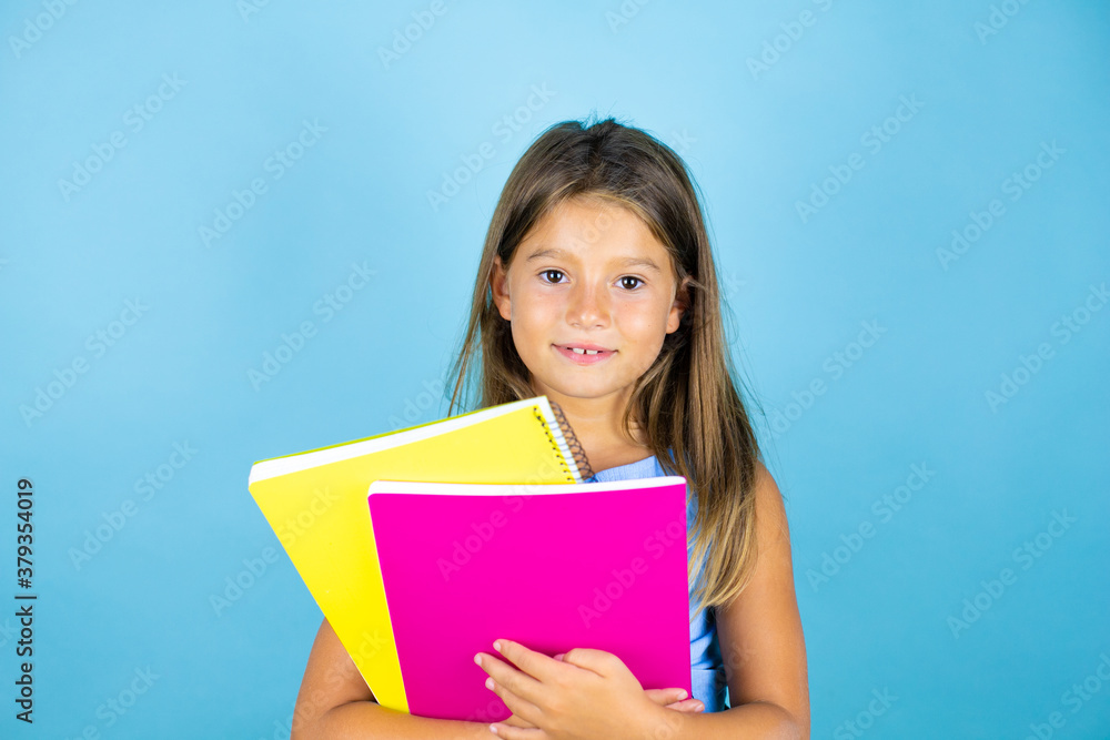 Young beautiful student child girl holding notebook over isolated blue background smiling and looking at the camera Young beautiful student child girl holding notebook over isolated blue background smiling and looking at the camera