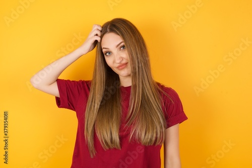 Beautiful Young beautiful caucasian girl wearing red t-shirt over isolated yellow background being confused and wonders about something. Holding hand on her head, uncertain with doubt. Pensive concept