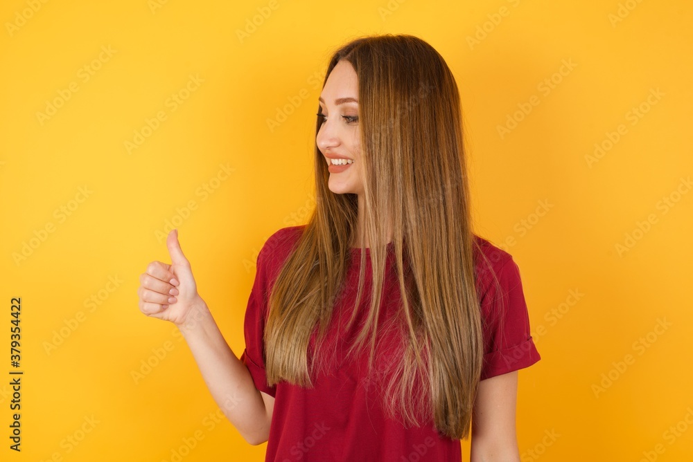 Beautiful Young beautiful caucasian girl wearing red t-shirt over isolated yellow background Looking proud, smiling doing thumbs up gesture to the side. Good job! Beautiful Young beautiful caucasian girl wearing red t-shirt over isolated yellow background Looking proud, smiling doing thumbs up gesture to the side. Good job!