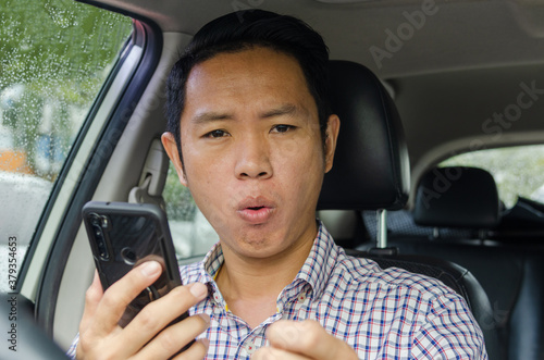 Asian man wearing a plaid shirt looks at smart phone in a car.