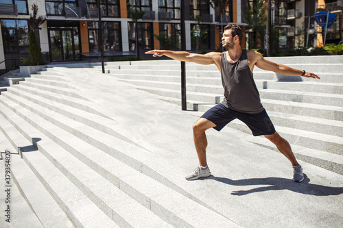 Young man exercising outside. Guy standing in yoga position alone at steps and stretching hands forward and backward. Take care of body. Urban training. Wonderful wellbeing. © Vlad