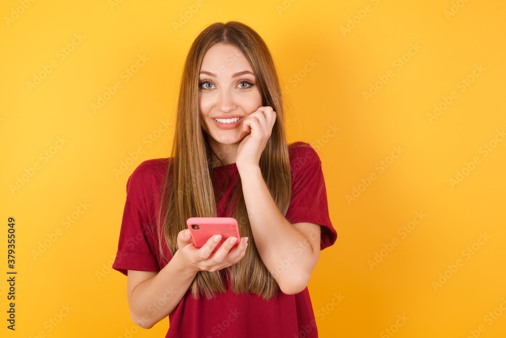 Beautiful Young beautiful caucasian girl wearing red t-shirt over isolated yellow wall poses with mobile phone device, types text message on modern smartphone, watches funny video during free time, Beautiful Young beautiful caucasian girl wearing red t-shirt over isolated yellow wall poses with mobile phone device, types text message on modern smartphone, watches funny video during free time,
