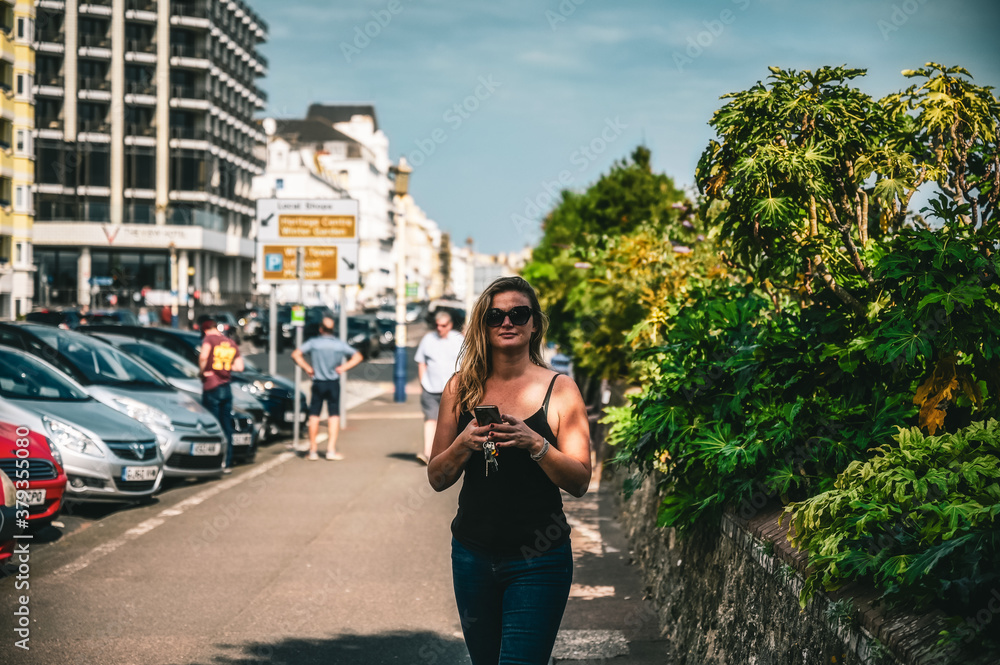 Lovely young blonde on seaside promenade looking at camera with phone in her hand