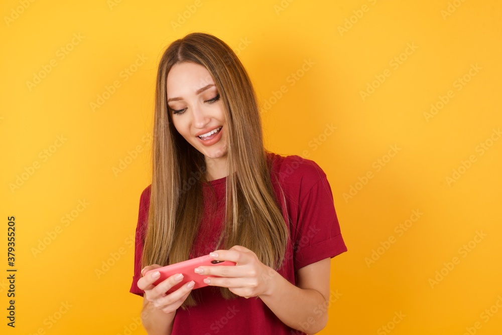 Close-up portrait of Beautiful Young beautiful caucasian girl wearing red t-shirt over isolated yellow background holding in hands cell playing video games or chatting