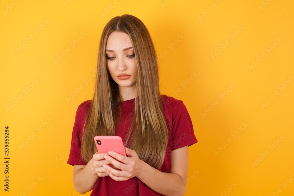 Excited Beautiful Young beautiful caucasian girl wearing red t-shirt over isolated yellow background winking and eye hold smart phone use read social network news Excited Beautiful Young beautiful caucasian girl wearing red t-shirt over isolated yellow background winking and eye hold smart phone use read social network news