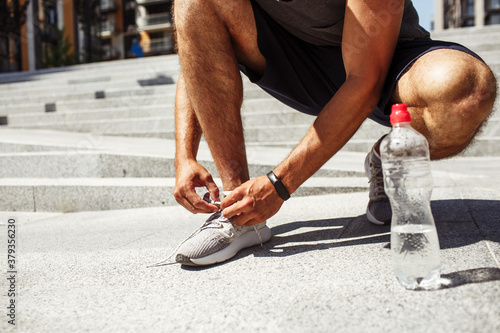 Young man exercising outside. Cut low view of man tie laces of shoes or sneakers. Almost empty water bottle besides on asphalt. Sporty athlete getting ready for exercising, training or having workout.