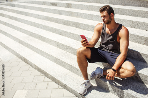 Young man exercising outside. Guy sit and rest on steps after hard workout. Hold smartphone in hand and look on it. Sexy handsome athlete posing. Modern technologies and devices.