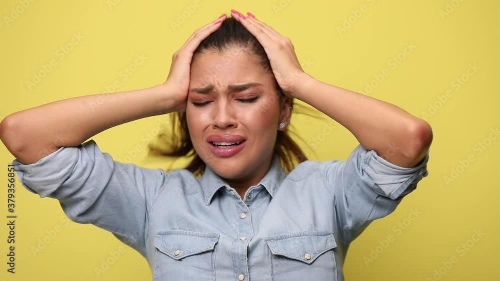 sad casual girl in blue denim shirt holding hands on head, nodding, crying and covering face with hands, refusing on yellow background sad casual girl in blue denim shirt holding hands on head, nodding, crying and covering face with hands, refusing on yellow background