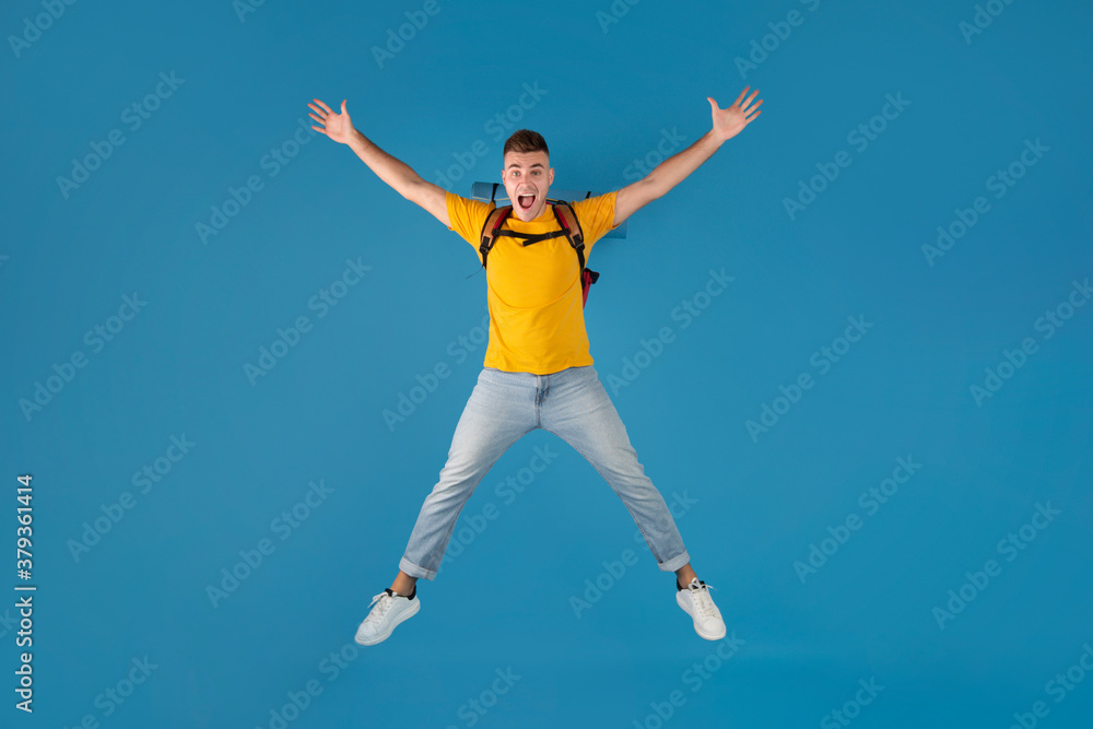 Handsome millennial guy with backpack jumping in excitement over blue studio background