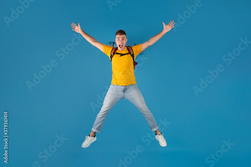 Handsome millennial guy with backpack jumping in excitement over blue studio background © Prostock-studio