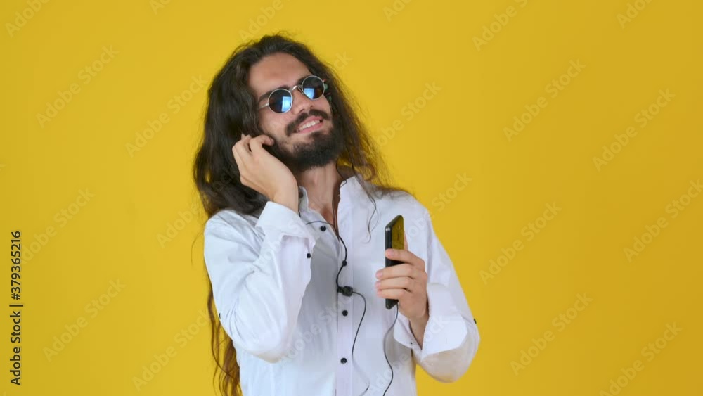A cute guy with long hair and a white shirt and sunglasses listens to music and dances on a yellow background. People lifestyle concept. A cute guy with long hair and a white shirt and sunglasses listens to music and dances on a yellow background. People lifestyle concept.