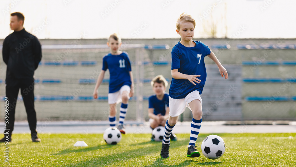 Group of School Kids with Young Coach Kicking Soccer Balls on Grass Sports Pitch. Junior Footballers in Blue Shirts on Training. Horizontal Image of Soccer Club Practice