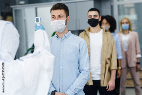Man in protective suit measures temperature of workers in protective masks in office interior, stopping spread of covid-19