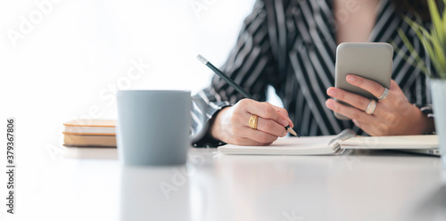 Cropped shot of female hand writing and using smartphone at office.