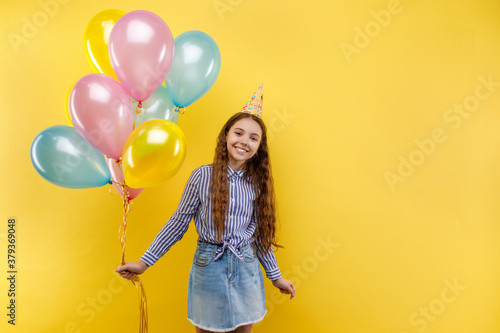 Young girl in birthday party with colorful inflatable ballons isolated on a yellow background, little beautiful girl wearing birthday cap, smiling with happy face. Copy space