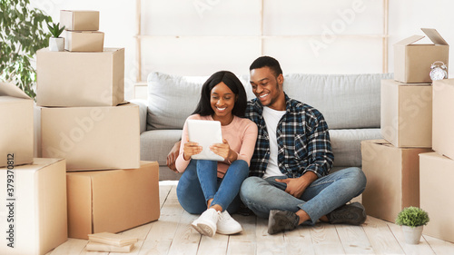 Cheerful black couple with tablet computer sitting on floor among cardboard boxes on relocation day
