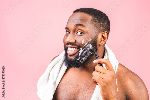 African American man smears shaving cream on face by shaving brush. Male hygiene. Isolated on pink background. Studio portrait.