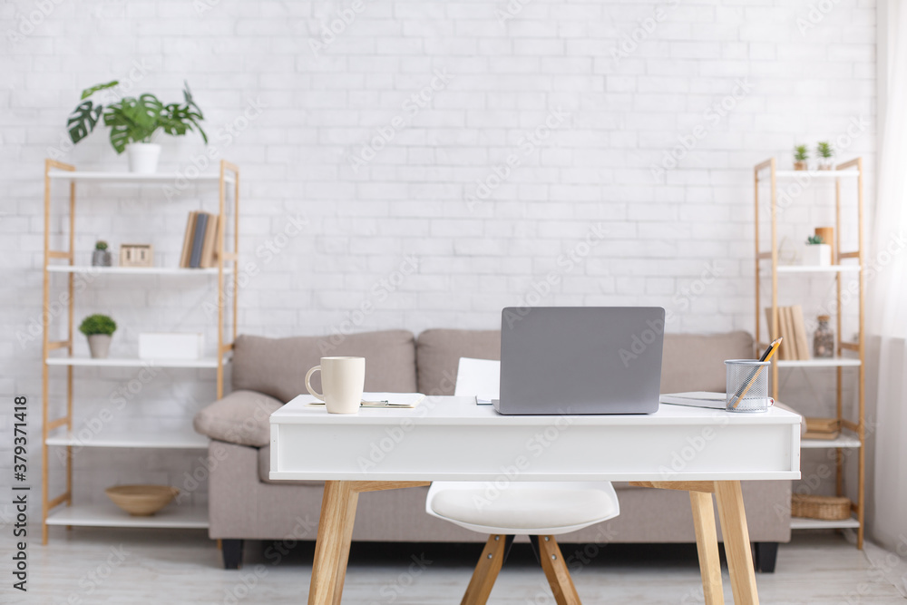 Fototapeta premium Remote workplace or home office during coronavirus outbreak. Sofa, chair, table with laptop, cup and notepad in living room interior