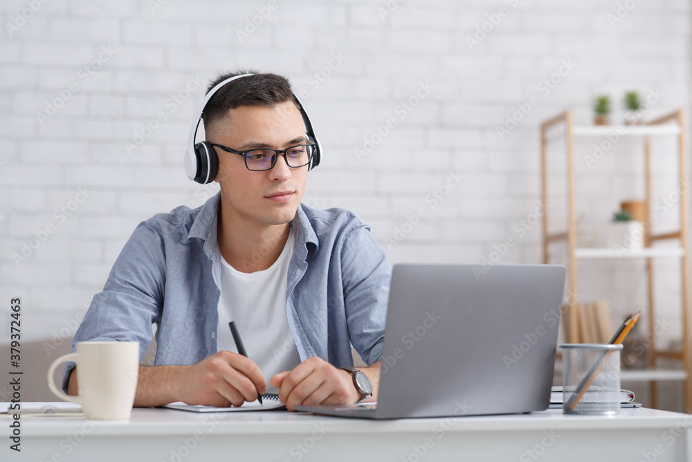 Modern student or pupil studies at home. Serious guy with glasses and headphones makes notes in notebook and looks at laptop, listening lecture Modern student or pupil studies at home. Serious guy with glasses and headphones makes notes in notebook and looks at laptop, listening lecture
