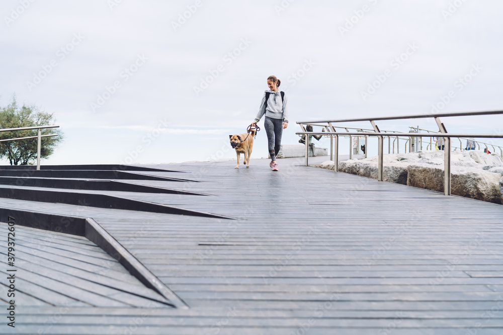 Carefree Caucasian female traveller 30s enjoying vacations time for rest with morgel dog, youthful woman with doggie on leash strolling during coastline promenade for walking pet at boardwalk