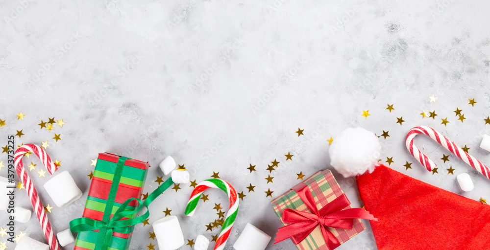 Christmas composition. Gifts, caramel cane, marshmellow, santa hat on gray concrete background with sparkling stars. Concept of winter holidays, new year, Christmas. Top view. Copy space