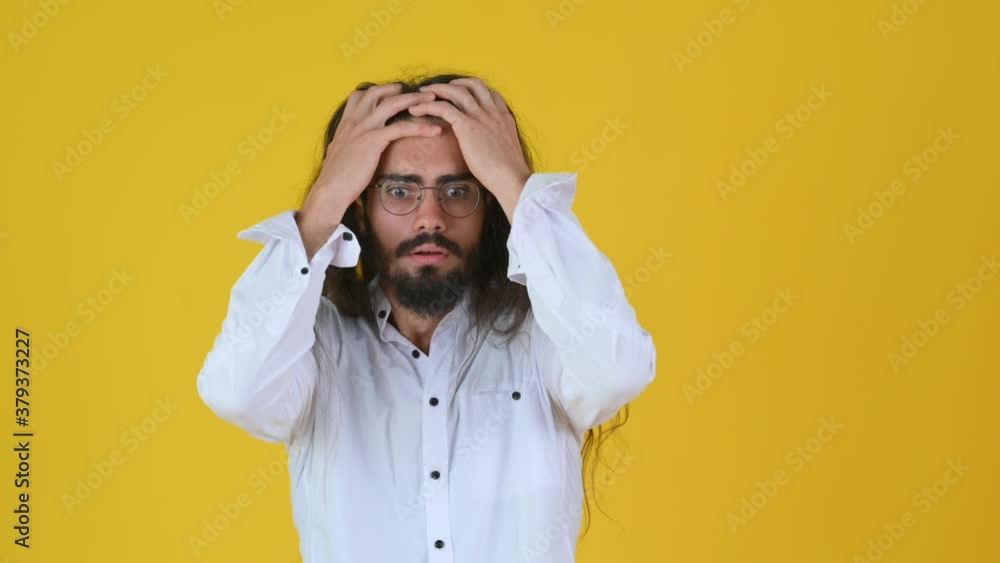 Shocked bearded young guy with long hair and white shirt surprised puts his hands to his head. Stressed and depressed guy isolated on yellow studio background Shocked bearded young guy with long hair and white shirt surprised puts his hands to his head. Stressed and depressed guy isolated on yellow studio background