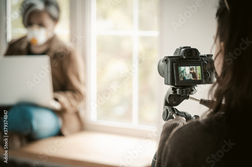Backstage of shooting. Selective focus on digital camera in foreground. Woman in face mask using laptop sitting near window on blurred background. Quarantine concept. © Svyatoslav Lypynskyy