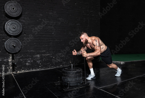 Young strong sweaty muscular fit man with big muscles pushing heavy weight plates on the prowler slider in the gym as hardcore cross workout training