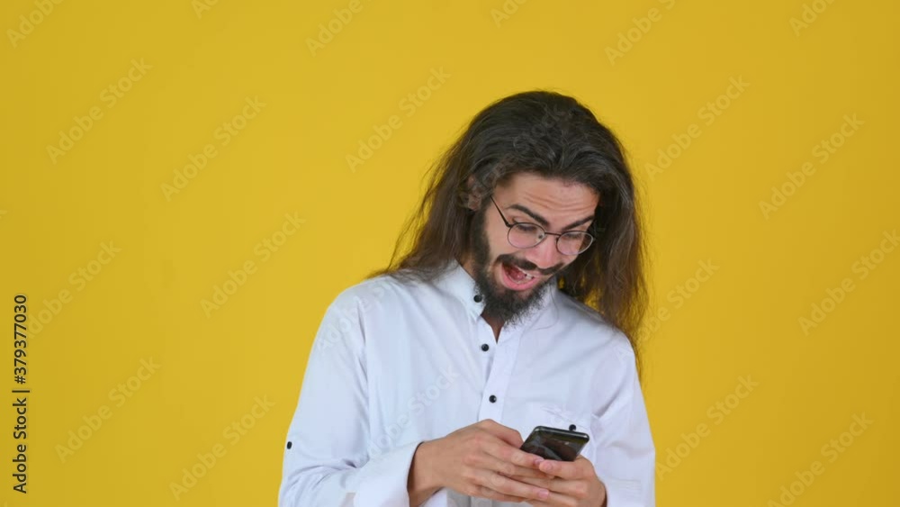 Handsome young guy with long hair in a white shirt looking at the phone screen and is surprised. People sincere emotions, lifestyle concept. Victory Handsome young guy with long hair in a white shirt looking at the phone screen and is surprised. People sincere emotions, lifestyle concept. Victory