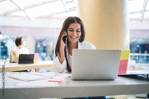 Cheerful caucasian female employee having positive conversation on mobile phone during job on laptop computer in office, smiling woman secretary assistant enjoying job process talking on smartphone