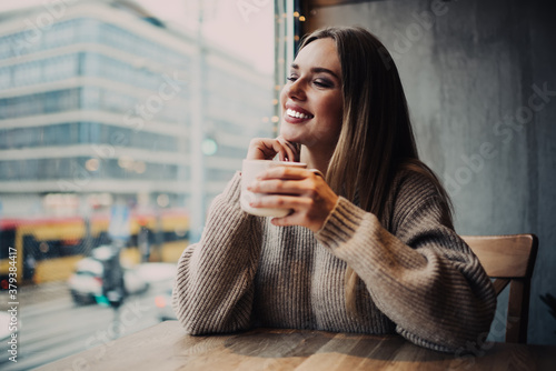 Cheerful hipster girl 20s with perfect veneers laughing during resting time in cafe interior, sincerely female customer with tea cup for warming smiling during cafeteria recreation on weekend photo