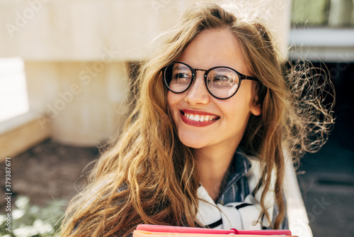 Closeup portrait of a smiling young student woman wearing transparent eyeglasses standing next to the college campus and carrying lots of books and folders on a sunny day. photo