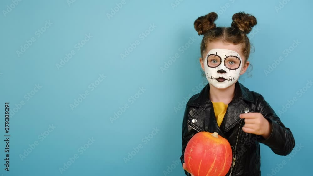 Cute preschooler girl point finger at orange pumpkin, wears frightening makeup and black leather jacket, looks at camera, isolated on blue studio background with copy space, celebrates Halloween Cute preschooler girl point finger at orange pumpkin, wears frightening makeup and black leather jacket, looks at camera, isolated on blue studio background with copy space, celebrates Halloween