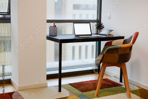 Office with a work table with a laptop, telephone, and pens, near a window, with a chair and a colorful carpet.