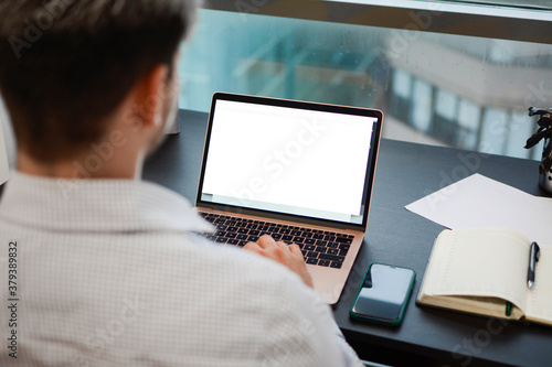 A man working on his laptop at his desk in front of the window.