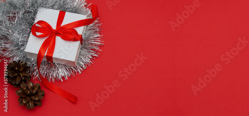 Christmas banner. The white gift box is tied with a red bow with Christmas tree branches, pine cones and silver mesh on a red background. Close-up, copy of the space.