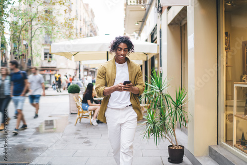 Handsome male tourist in casual trendy wear walking on urban settings holding mobile phone using navigation app, smiling hipster guy checking notification on smartphone about income text messages