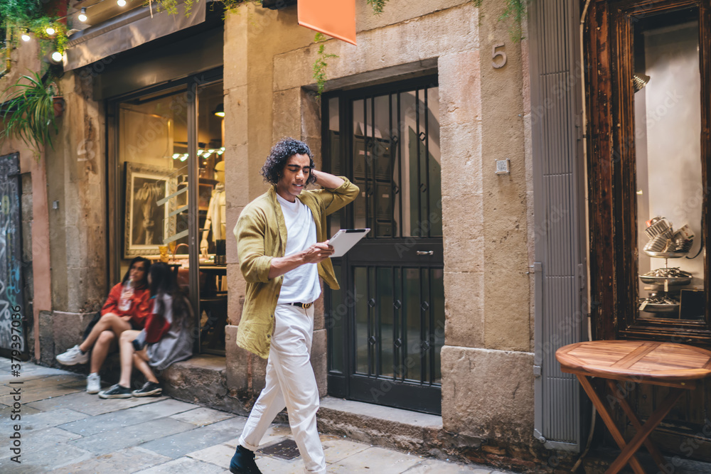 Handsome curly hipster guy in casual wear walking on urban settings using application on digital tablet for searching places to eat, smiling young male strolling and sending mails on touchpad outdoors