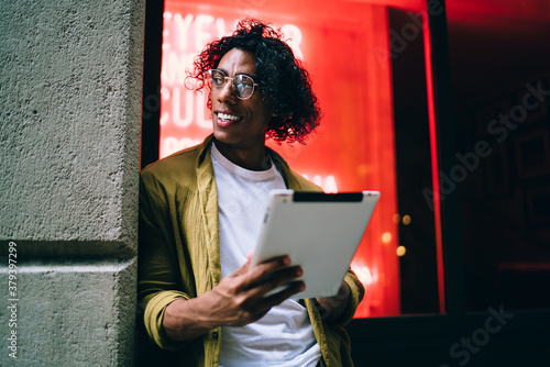 Smiling curly hipster guy in eyewear standing on neon light street with digital tablet share contetnt in vlog,handsome man influencer using touchpad for blogging and share media and multimedia content