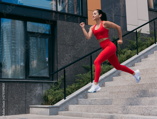 Athletic fit young woman with brown ponytail in red sports top and leggins running. Girl in white sneakers jumping down from stairs. Urban sport concept. Apartment block on background.