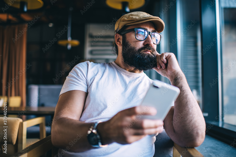 Pensive caucasian bearded male using mobile phone spending free time in coffee shop dreaming about plans and ideas, handsome mature man in eyewear pondering and resting with smartphone in cafeteria Pensive caucasian bearded male using mobile phone spending free time in coffee shop dreaming about plans and ideas, handsome mature man in eyewear pondering and resting with smartphone in cafeteria