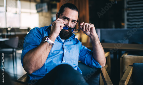 Portrait of handsome male 40 years in casual wear resting in cafe interior during free time, confident mature bearded man taking off spectacles looking at camera sitting indoors concentrated on ideas