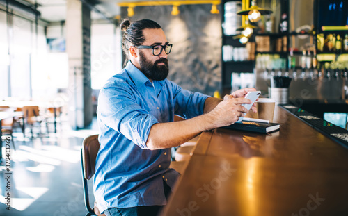 Serious adult hipster male using smartphone while sitting at bar counter of cafe in daytime